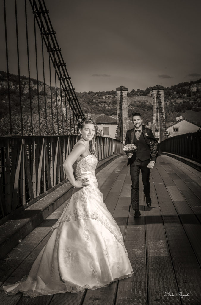 photo en noir et blanc d'un couple de jeune marié sur le pont de la ville de Cajar en Aveyron la riviére du lot passe en dessous la mariée est au premier plan avec une robe blanche le marié en arrière plan courre vers la mariée avec le bouquet de fleurs du mariage